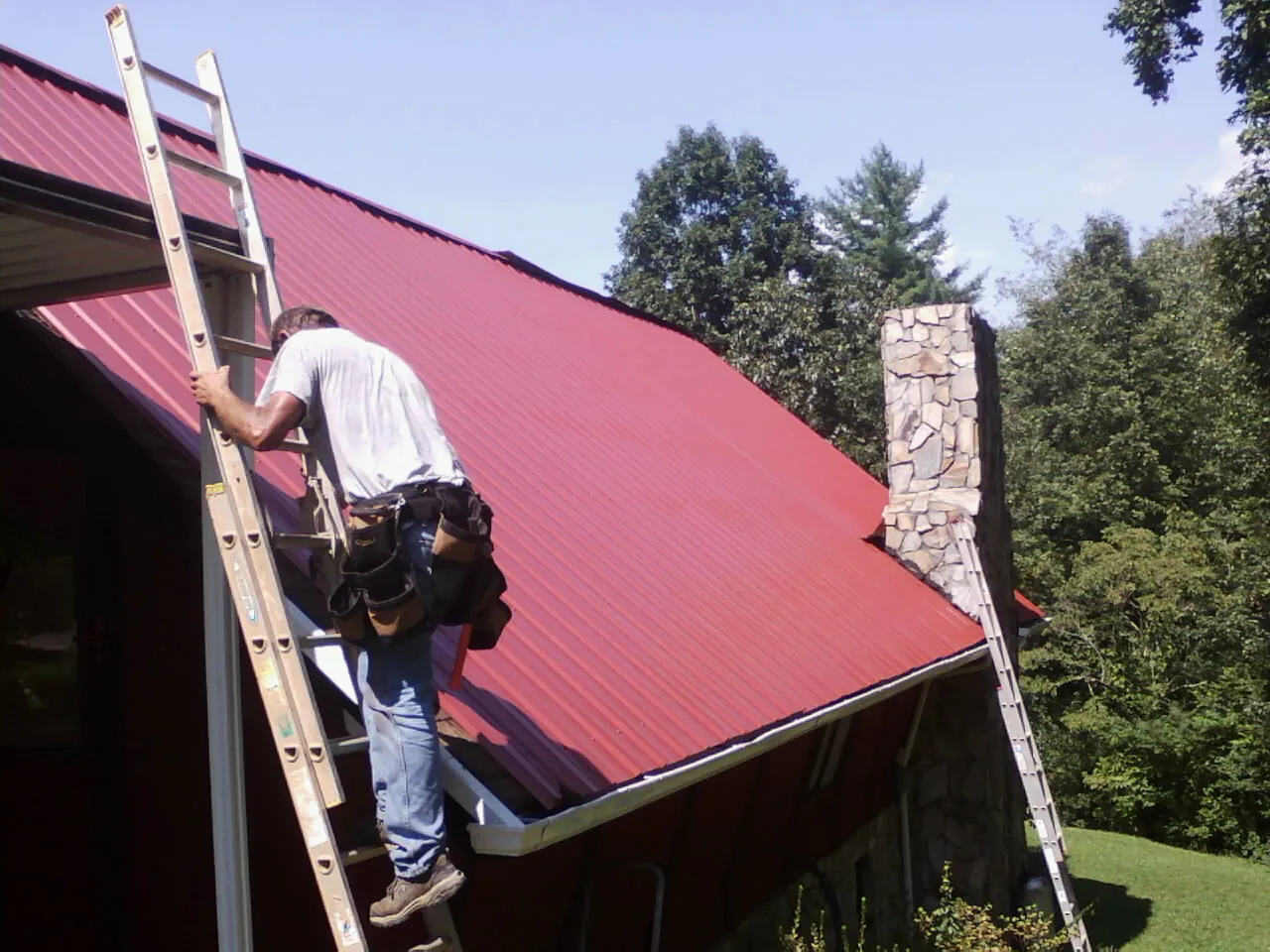 Roofer on a ladder beside a red metal roof and stone chimney — Soffit & Fascia Repair by Roofing Brothers in Bird Island